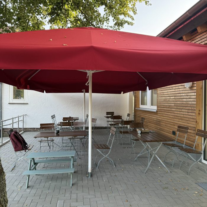 Auf einer gepflasterten Terasse stehen zwei große rote Schirme und darunter einige Tische mit Stühlen.