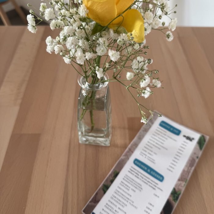 Auf einem Tisch steht eine kleine gläserne Vase mit einer gelben Rose darin. daneben liegt eine Speisekarte.