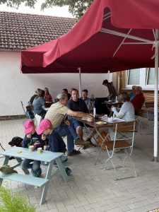 Auf einer Terasse sitzen einige Menschen und trinken Kaffee. Im Vordergrund sitzen zwei Kinder an einem Spieltisch.