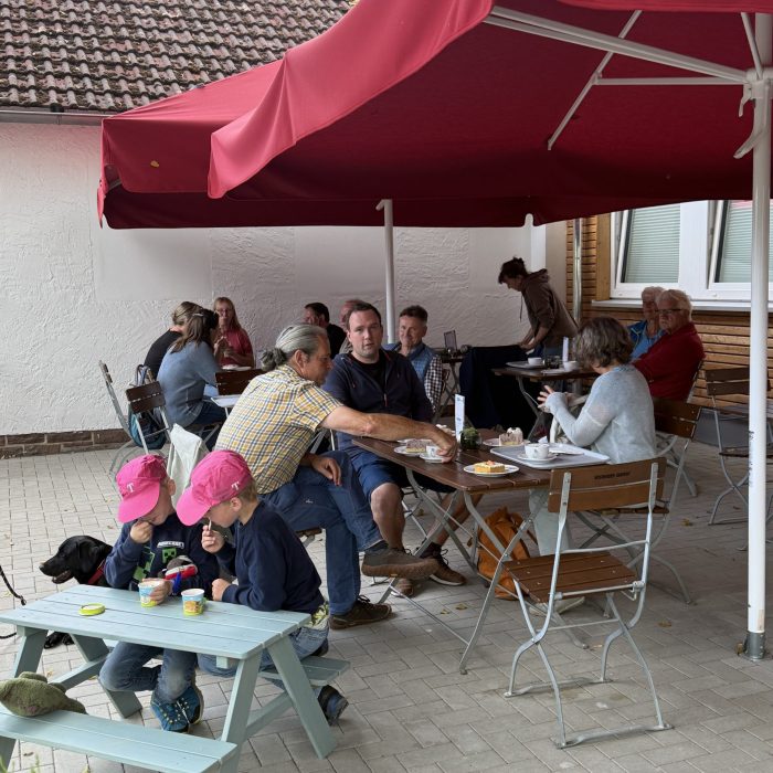 Auf einer Terasse sitzen einige Menschen und trinken Kaffee. Im Vordergrund sitzen zwei Kinder an einem Spieltisch.