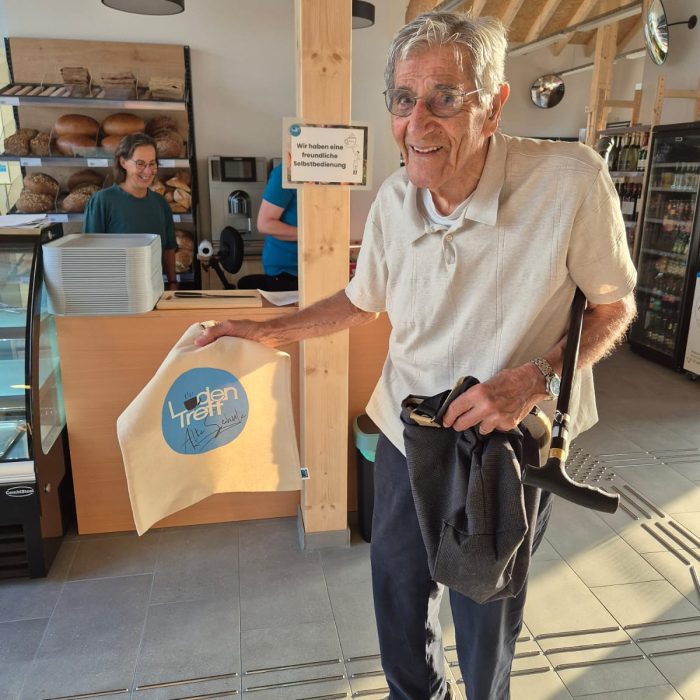 Ein älterer Herr mit Brille und Gehstock unterm Arm hält eine Stofftasche mit dem LadenTreff Logo hoch.