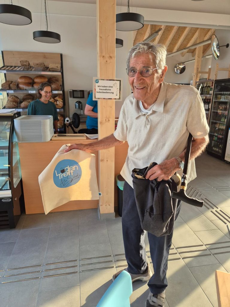 Ein älterer Herr mit Brille und Gehstock unterm Arm hält eine Stofftasche mit dem LadenTreff Logo hoch.